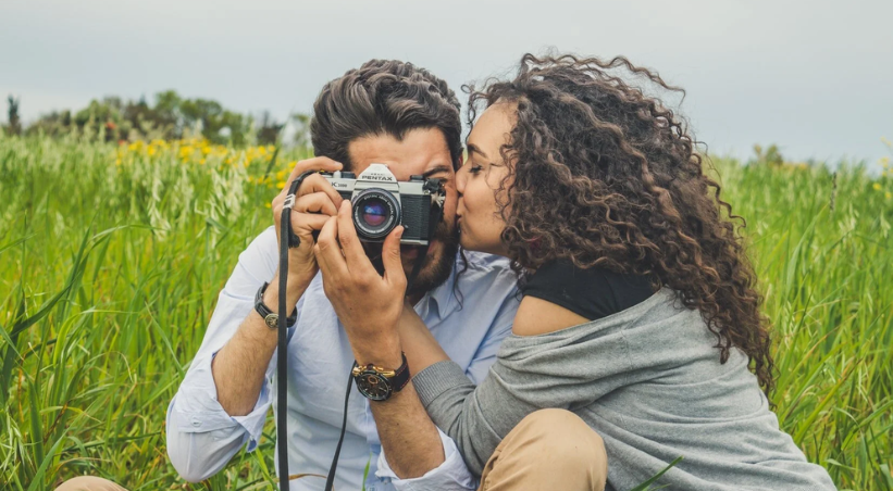 Young Indian couple sitting together in a green field, man holding a vintage camera while woman leans close to him looking at the camera display, showing shared interest and intimate connection in an outdoor setting.