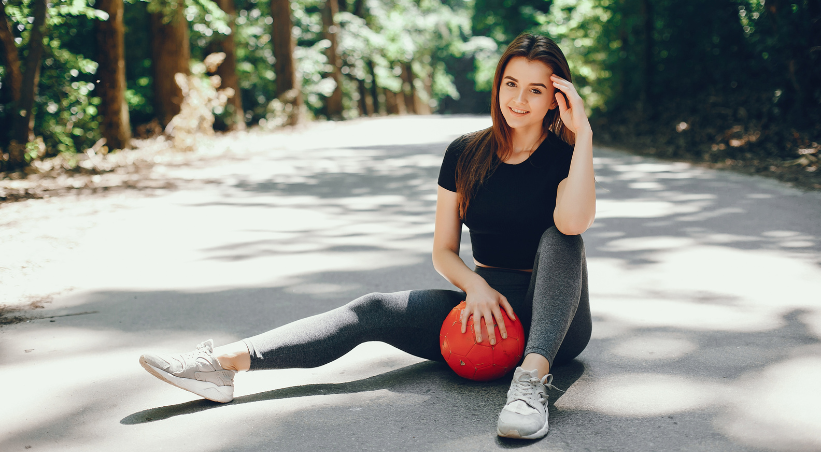 Smiling working woman in black t-shirt and gray leggings sitting on outdoor path with red medicine ball, representing active lifestyle and fitness routine.