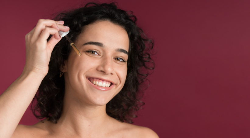 Woman applying serum for oily skin with dropper, smiling against burgundy background.