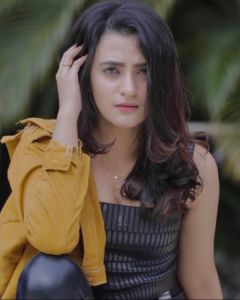 A woman with long dark hair wearing a black outfit and a mustard-yellow jacket, posing outdoors with a calm and confident expression.