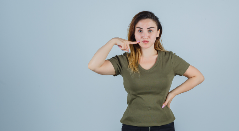 Young woman pointing to her cheek demonstrating facial exercise technique to reduce face fat at home.
