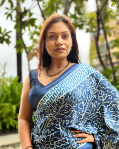 A woman wearing a blue patterned saree with a bindi, standing outdoors amid greenery with a confident and composed expression.