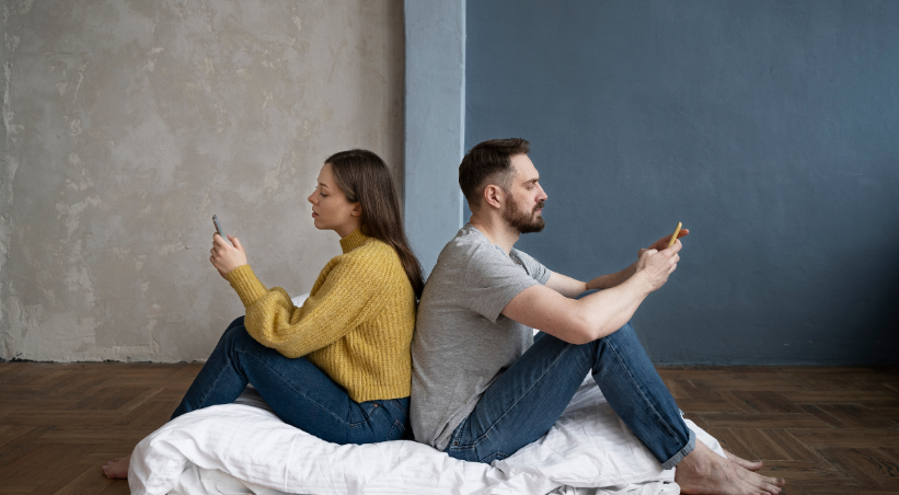 Young couple in long-distance relationship sitting back-to-back on bed using smartphones to video call and stay connected despite physical distance.