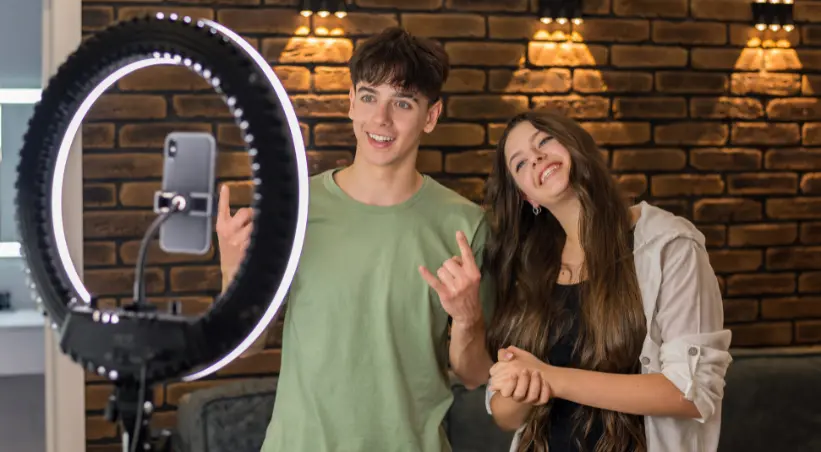 Two young content creators smiling beside a ring light and smartphone, recording an Instagram Reel in a cozy brick-wall studio.